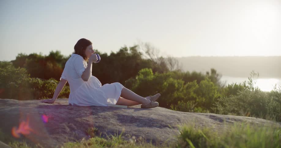 A woman is sitting on a large rock and drinking coffee by the lake