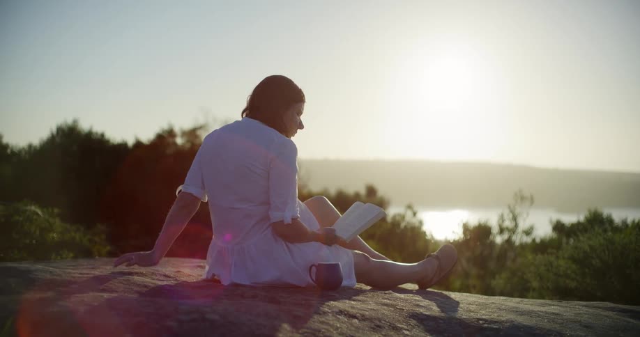 Pregnant woman in white dress sitting reading a book on a rock outdoors with a lake and mountains in the background