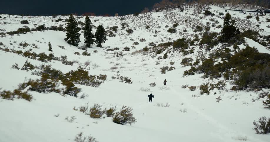 Two people hike through heavy snow down a hill toward a lake with snowcovered mountains rising on the opposite side under the midday sun