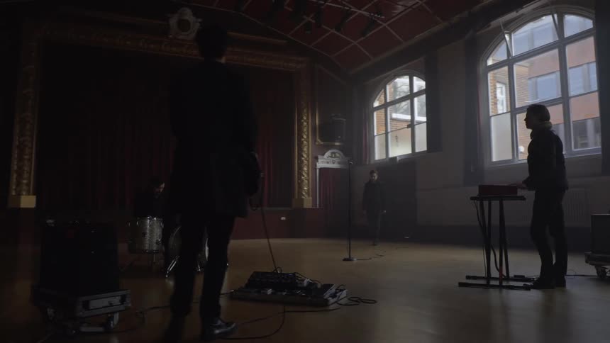 A singer walks in towards a microphone as a band performs in a large hall