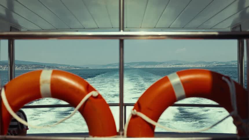 Looking out from the back of a moving boat two orange lifebuoys frame the view of the churning wake receding towards a distant hazy shoreline