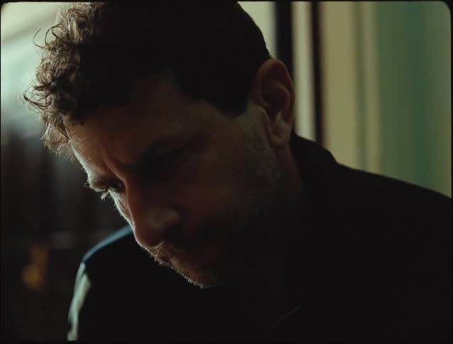 A man sits alone indoors looking down appearing thoughtful or sad