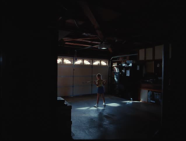 A young woman performs squats alone in a dimly lit garage illuminated by a beam of natural light