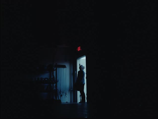 A lone person stands in a dark room silhouetted by light streaming in from an open exit door suggesting a moment of pause or decision
