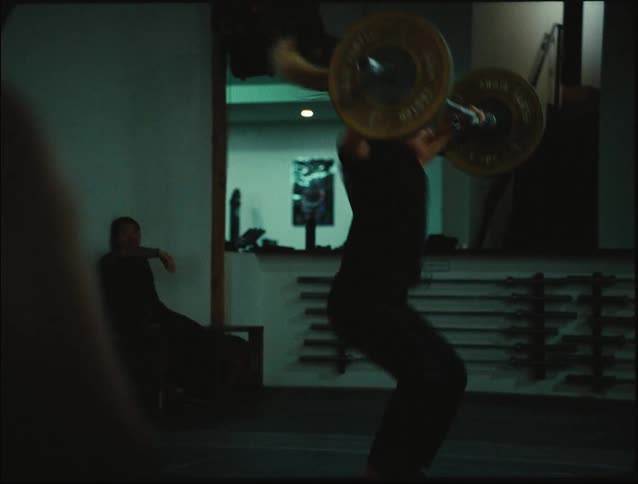 A woman is doing a snatch motion using free weights at the gym while one person is watching