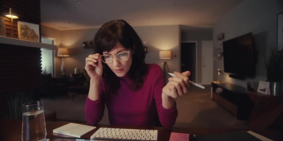 A woman in a maroon shirt sits at a desk puts on her glasses talks and uses a stylus on a device with a living area visible behind her