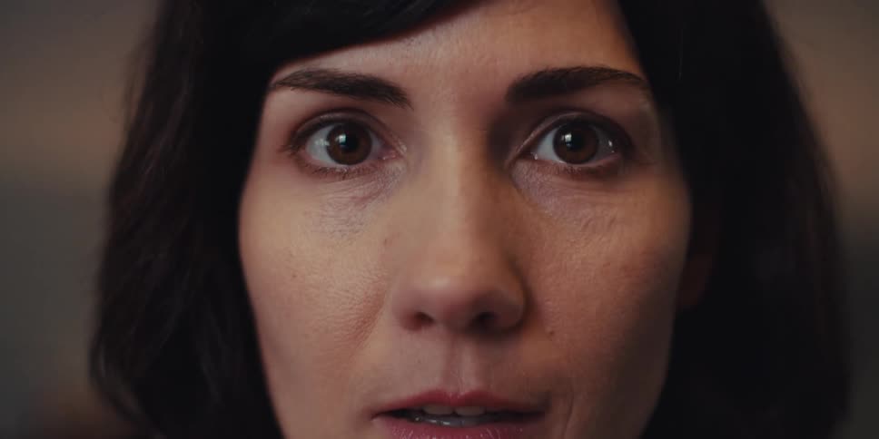 A woman with long black hair wearing a blazer types on a laptop while making small gestures and muttering to herself