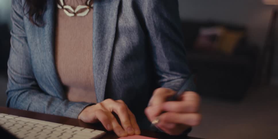 A woman in a professional setting writes on a sticky note while working at her desk then pauses to reflect with a thoughtful expression