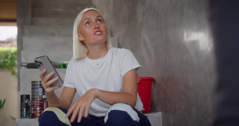A woman sitting on a stairway holding a phone and smiling as she animatedly converses with someone standing offcamera