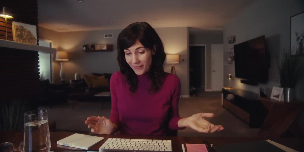 A woman in a maroon sweater smiles and types during a video call in a cozy dimly lit living room