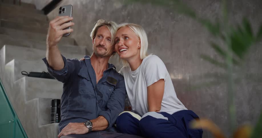 A smiling couple sits on a staircase taking a selfie during a video call