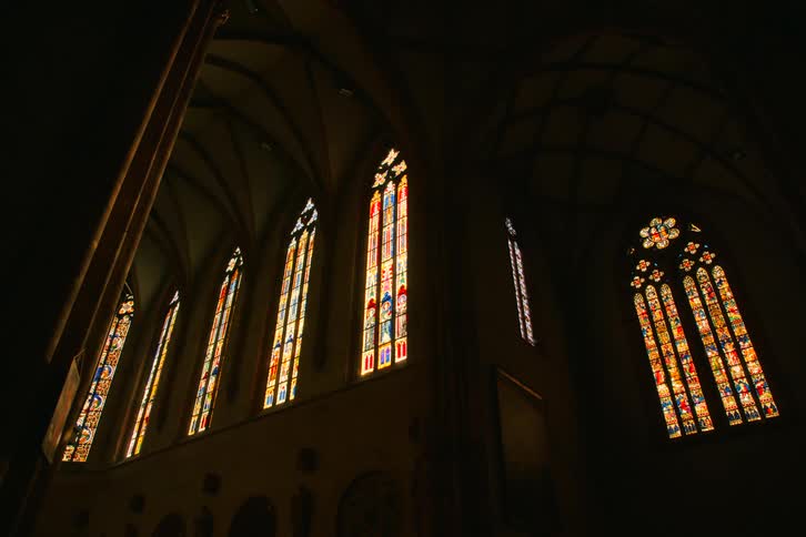 Sunlight illuminates tall ornate stained glass windows set within the higharched shadowy of a cathedral