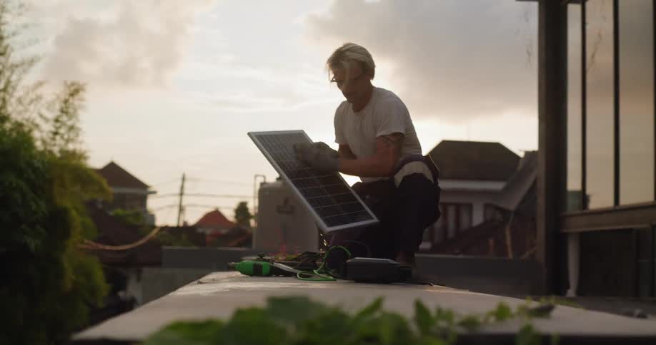 A man is shown working on a rooftop at sunset picking up and positioning a solar panel for installation