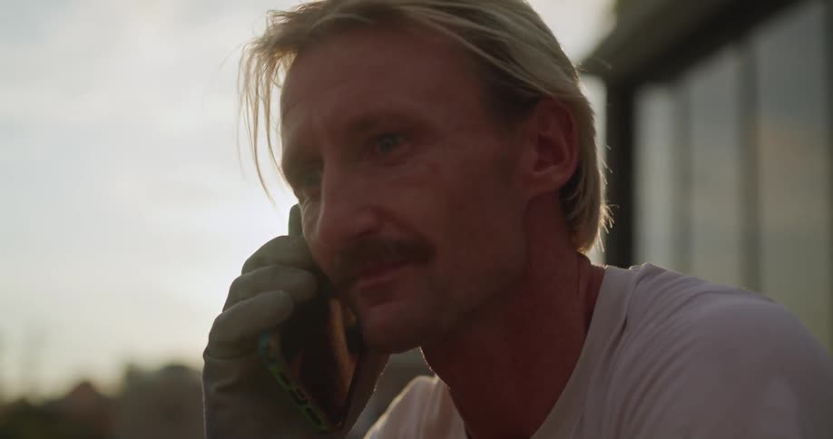 A man with a mustache talks on a cellphone outside during sunset looking thoughtful and slightly worn from work