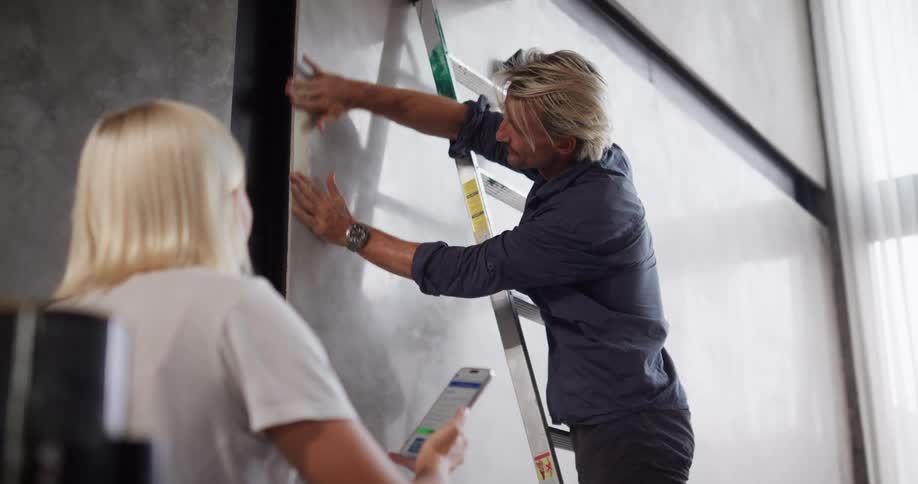 A man applies tape to a wall near a ladder while a woman observes with asmart phone in a modern interior
