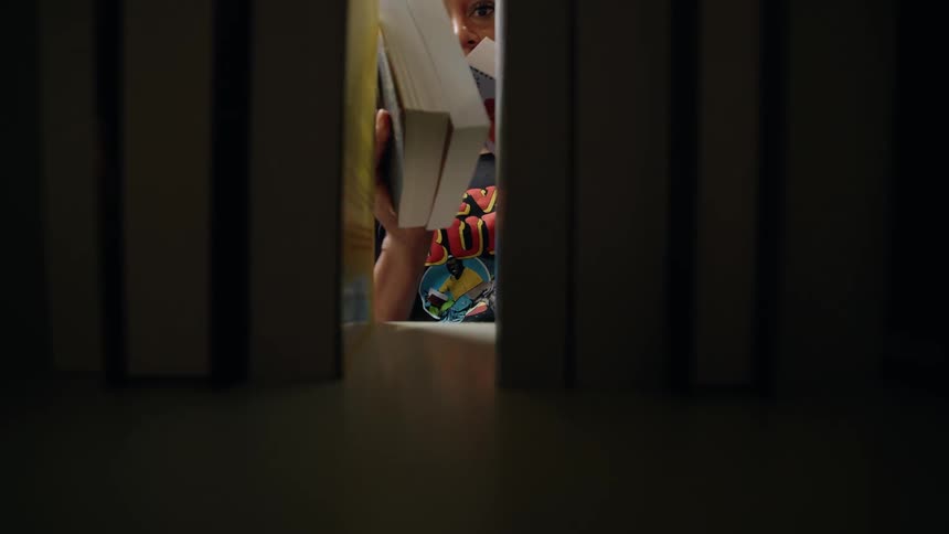 A person wearing a tshirt places books on a shelf as seen from a low angle between the books