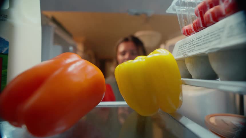 A woman opening the refrigerator to grab a bell pepper and then closing it