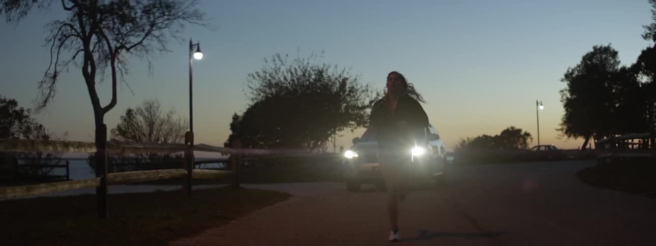 A car follows a woman jogging on a road at dusk