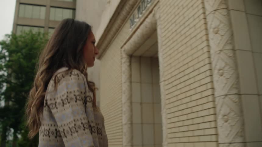 A woman wearing a patterned sweater and jeans walks up concrete steps and approaches a glass door to enter a building