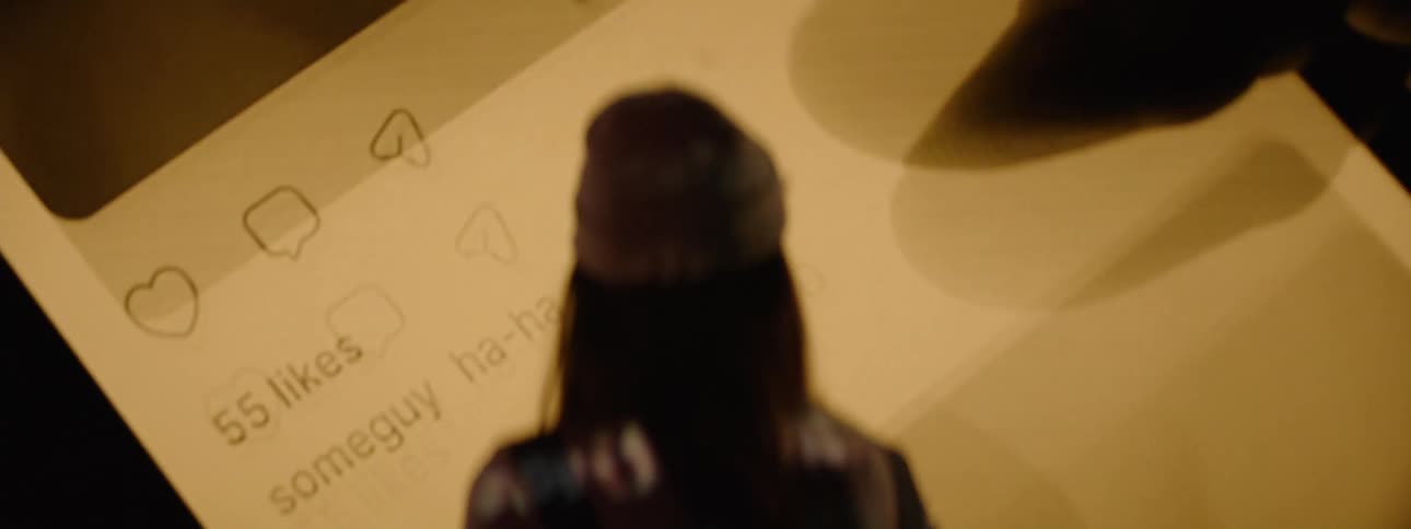 A woman wearing a beanie is standing in front of a large screen with the video of a person scrolling on their smart phone projected on it