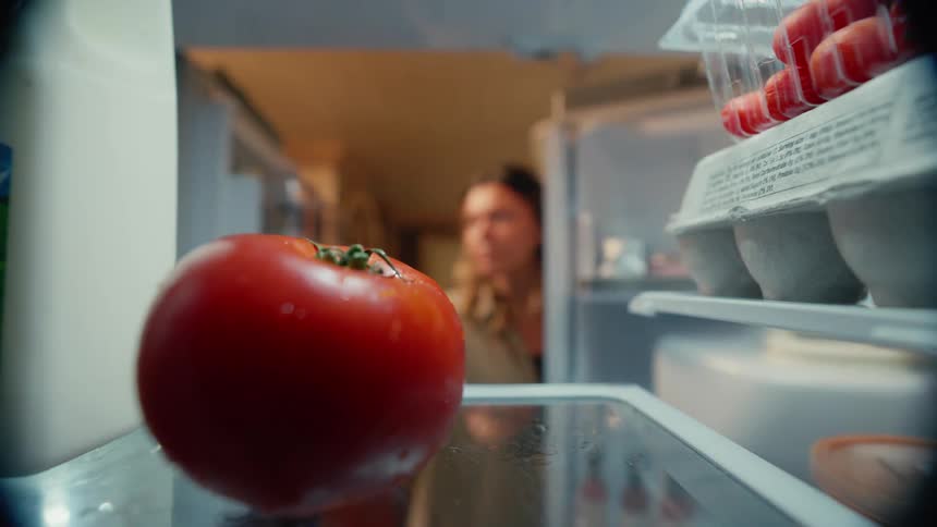 A woman opens a refrigerator and takes a tomato