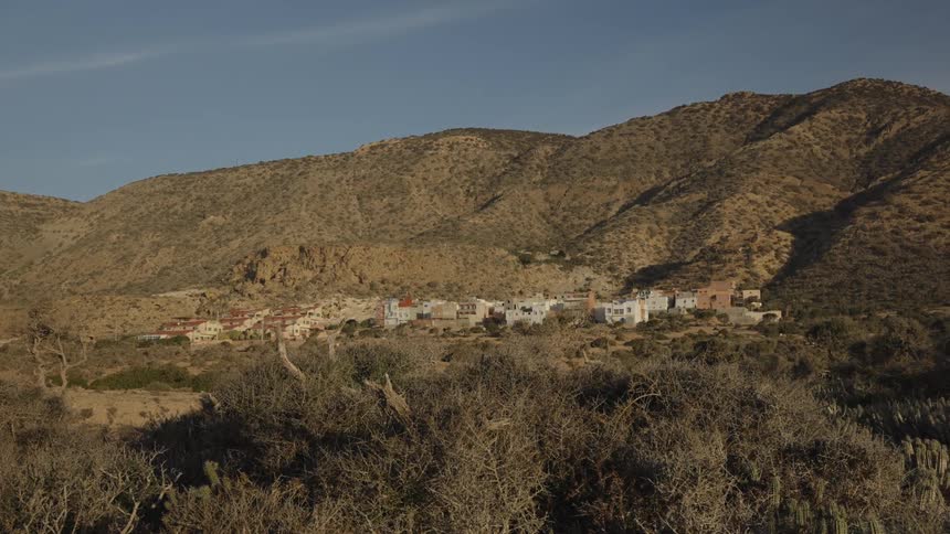 A small rural village sits nestled at the base of arid sunlit mountains under a clear blue sky