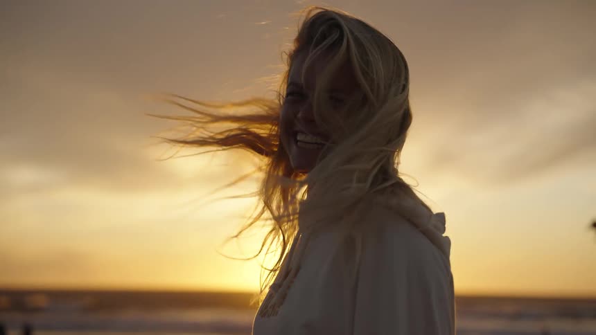 A woman in a grey sweatshirt with long blonde hair turns whipping her hair around as she laughs and faces the person filming her her silhouette framed against a low setting sun on the horizon over a calm ocean
