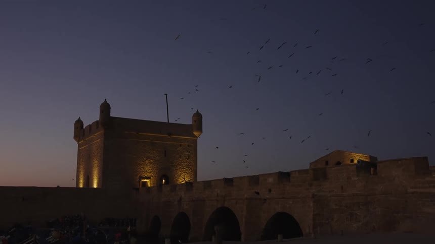 A historic stone fortress glows at dusk as birds soar across a twilight sky