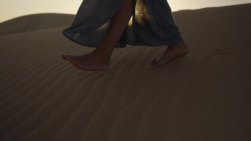 Legs of a lady walking barefooted in the desert at sunset leaving footprints