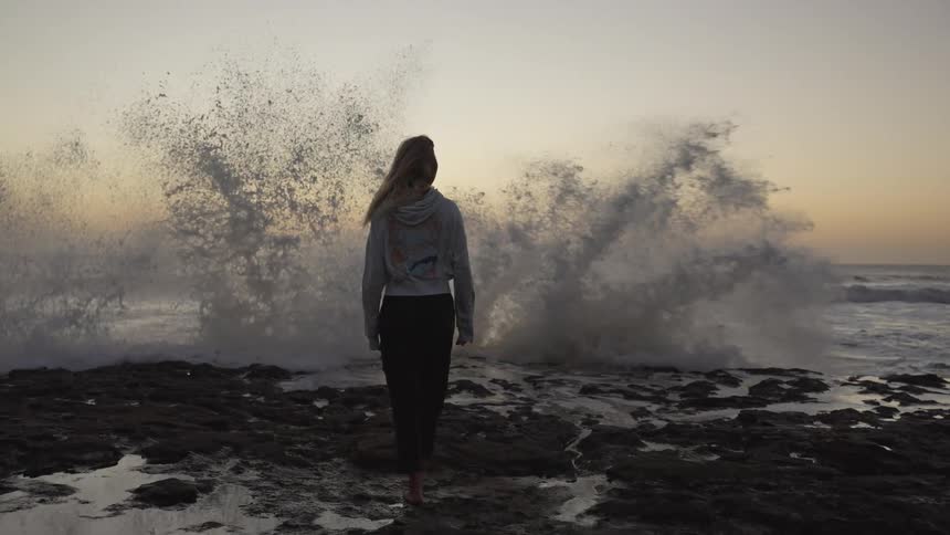 A woman stands barefoot on a rocky shore at sunset watching massive waves crash dramatically in front of her