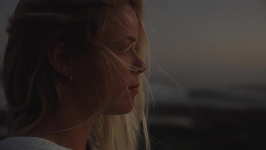 A woman with windblown hair gazes thoughtfully toward the ocean at sunset or sunrise