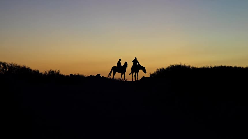 Silhouettes of two riders and their horses are seen on a ridge against a gradient sunset sky one horse turns and starts to walk away followed by the other