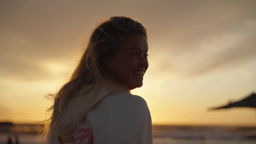 A woman turns and smiles at the camera while walking along a beach during a golden sunset