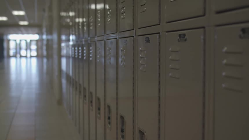 The camera moves slowly forward along a row of closed metal lockers in an empty school hallway with bright light visible from doors at the far end