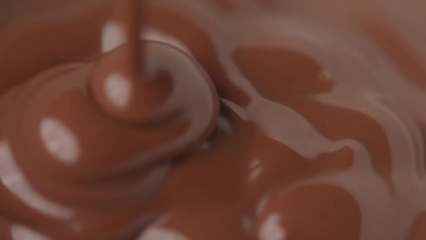 A thick stream of melted chocolate is poured on top of a pool of already melted chocolate bubbling and reflecting the light