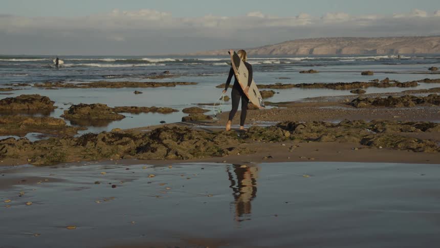 Female surfer carrying surfboard walking in low tide towards the beach