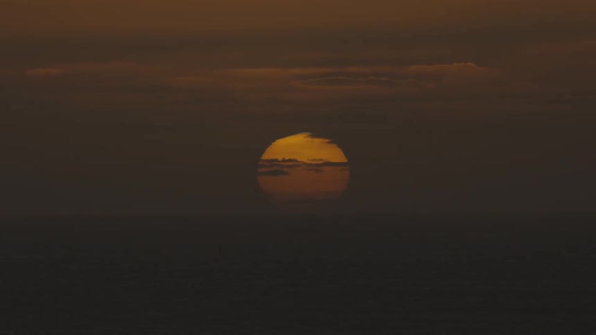 Setting sun glows through the clouds on the horizon over the water in a deep dark red sky