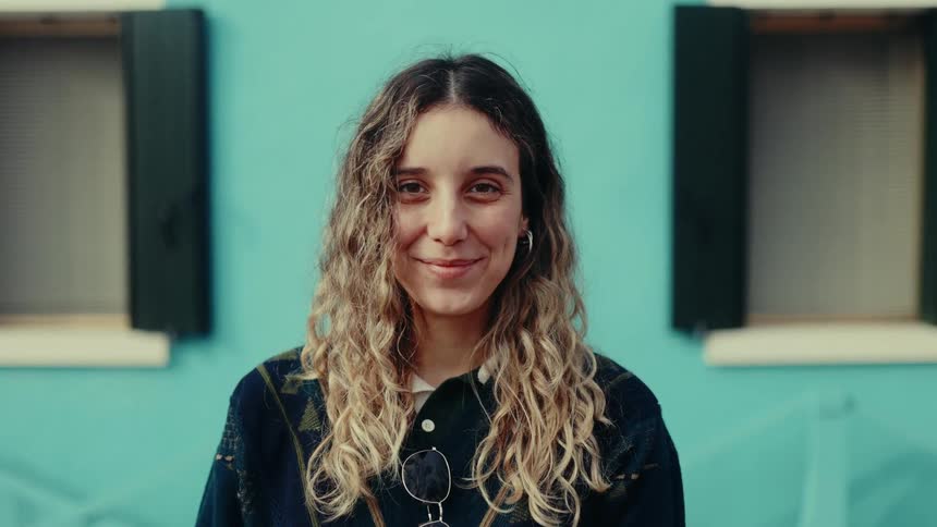 Curly haired woman with happy facial expression outdoors with a wall with windows in the background