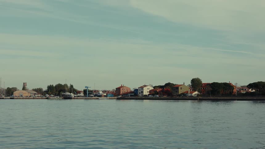 A view from the water shows a coastal town with various buildings and boats docked along the shoreline under a partly cloudy sky