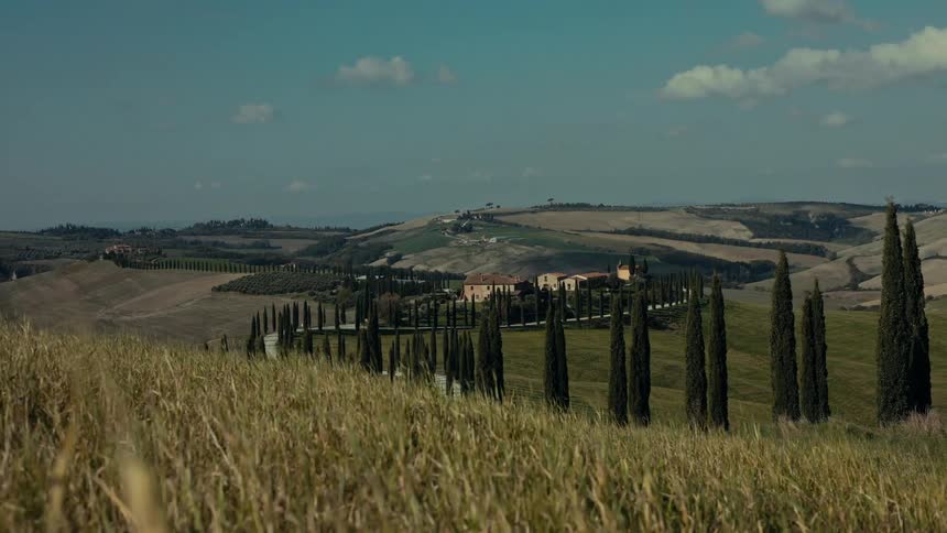 A wide shot frames a tuscan estate nestled among golden fields and lined with tall cypress trees beneath a clear sky