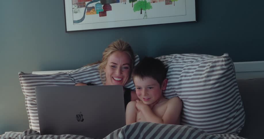 A woman and a young child are looking at a laptop screen while nestled together in a bed with striped linens