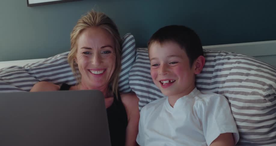 A mother and her young son are joyfully laughing together while watching something on a laptop as they relax in bed