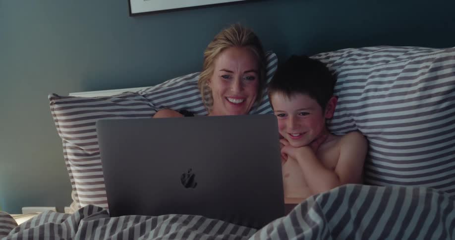 A mother and her son cuddle in bed while happily watching something together on a laptop