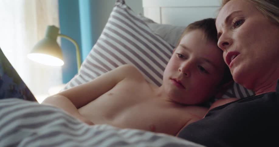 Mother reads book to her son at daytime with their backs on the bed