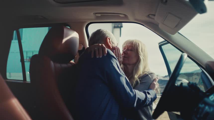 An older couple shares a tender kiss in a parked car during an emotional goodbye