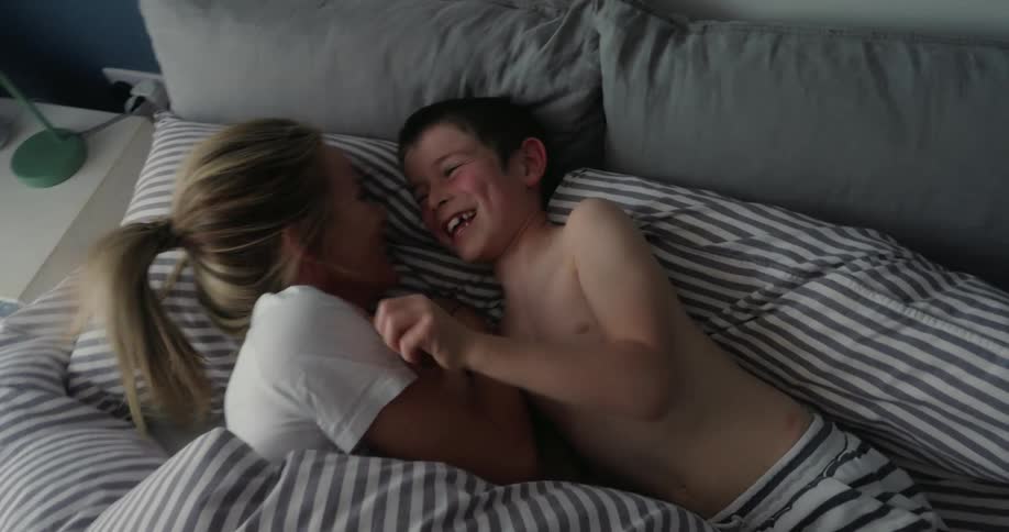 A woman and a young boy are playfully laughing and tickling each other in bed sharing a joyful moment