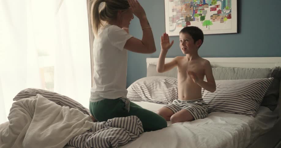 A woman and young boy sit on a bed playing a handclapping game in a warmly lit bedroom