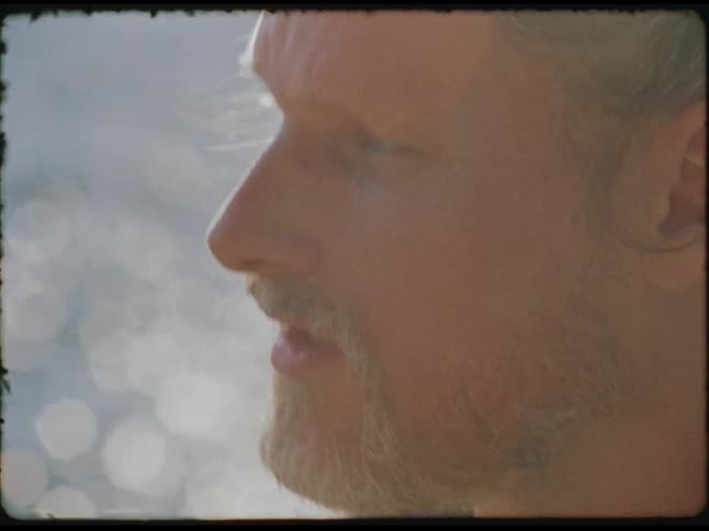 A bearded man is shown in closeup profile singing followed by a closeup of his hands expertly playing an acoustic guitar against a softly blurred sundappled background