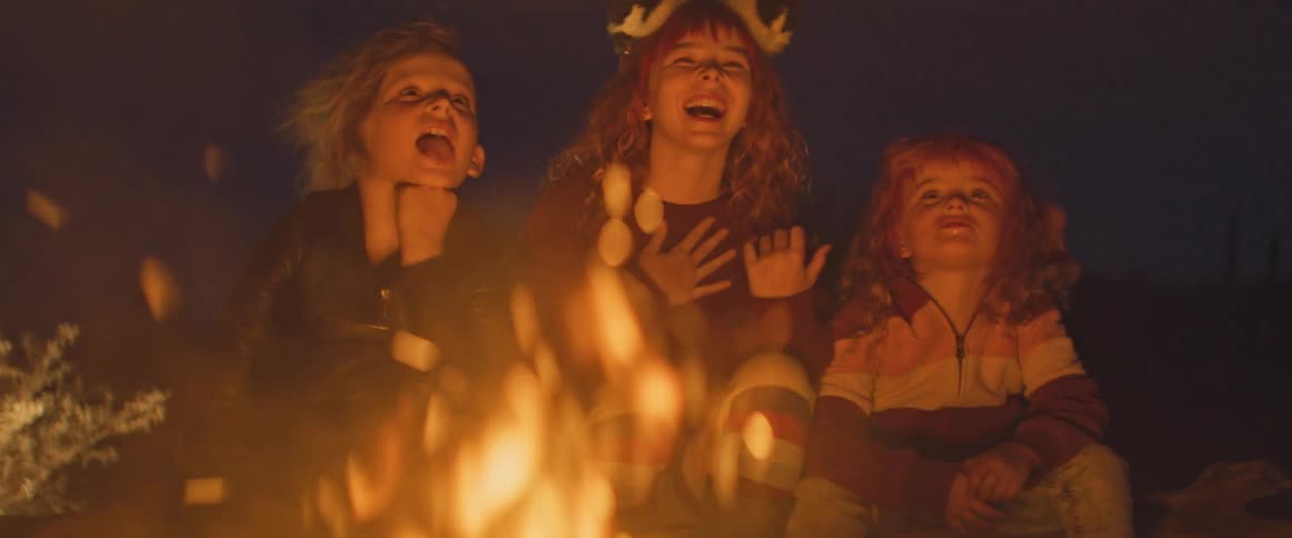 Three children are sitting around a fire at night laughing and enjoying themselves