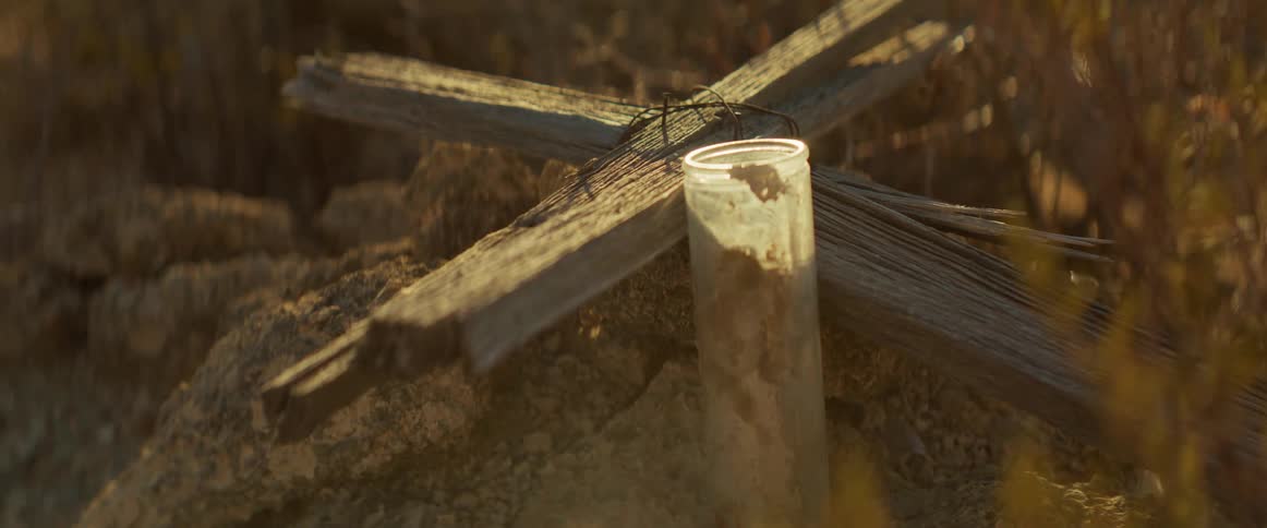 A weathered cross and candle rest in rocky terrain creating a somber reflective mood